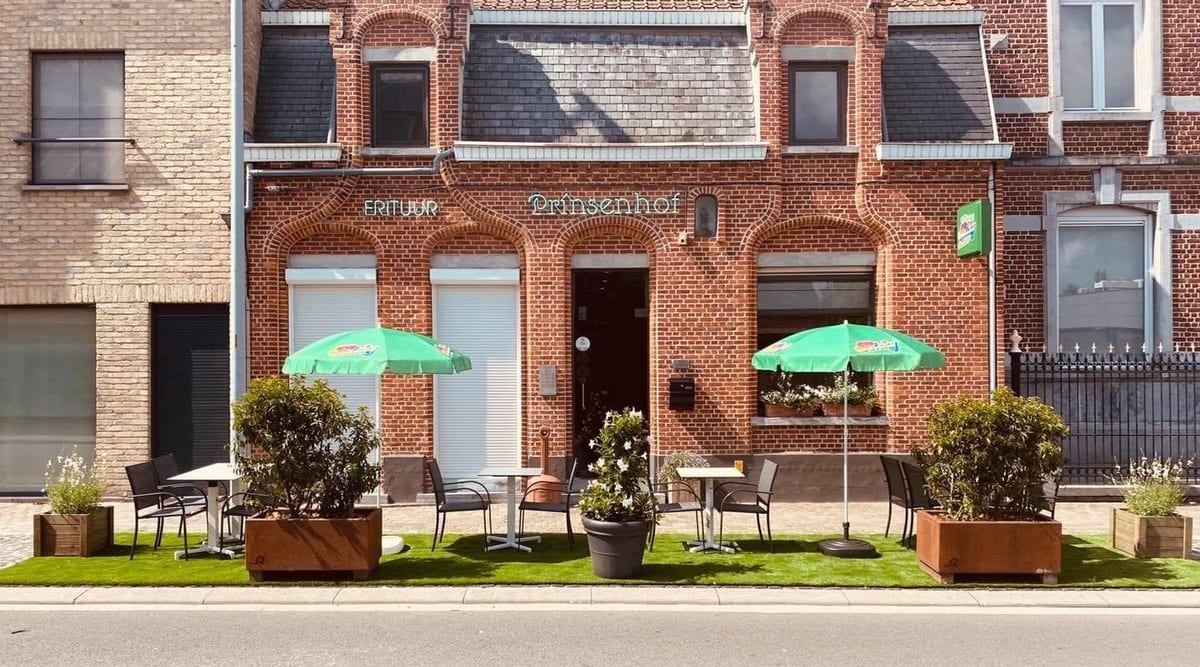 De rode bakstenen gevel van Frituur Prinsenhof met de neonnaam boven de boogramen, groene parasols en terrastafels op het kunstgras
